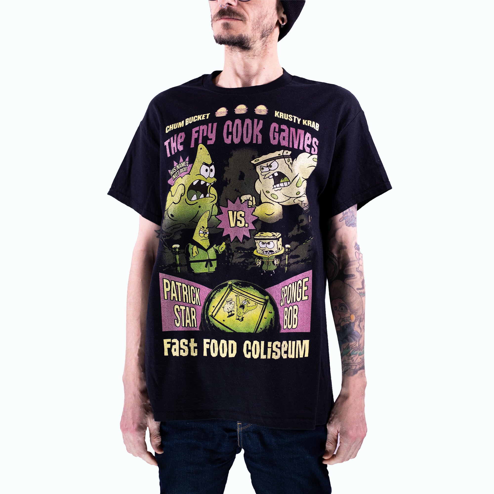 Male body fit shot of a Black t-shirt featuring a jumbo graphic of "The Fry Cook Games" from SpongeBob SquarePants. The design shows muscular versions of SpongeBob and Patrick facing off in a wrestling-style match, sponsored by the Krusty Krab and Chum Bucket. Text includes “His name’s not Rick!”, “Patrick Star vs. SpongeBob”, and “Fast Food Coliseum” with a green filtered wrestling ring at the bottom. The style is bold, distressed, and retro-inspired.